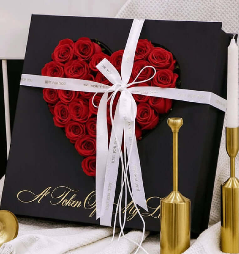 Heart-shaped arrangement of red roses in a black box with a white ribbon, placed on a textured surface with gold candle holders.