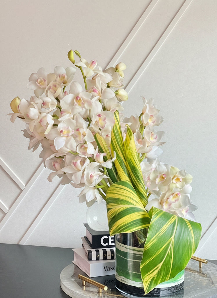 A modern floral arrangement featuring white Cymbidium orchids and green foliage in a clear glass vase.