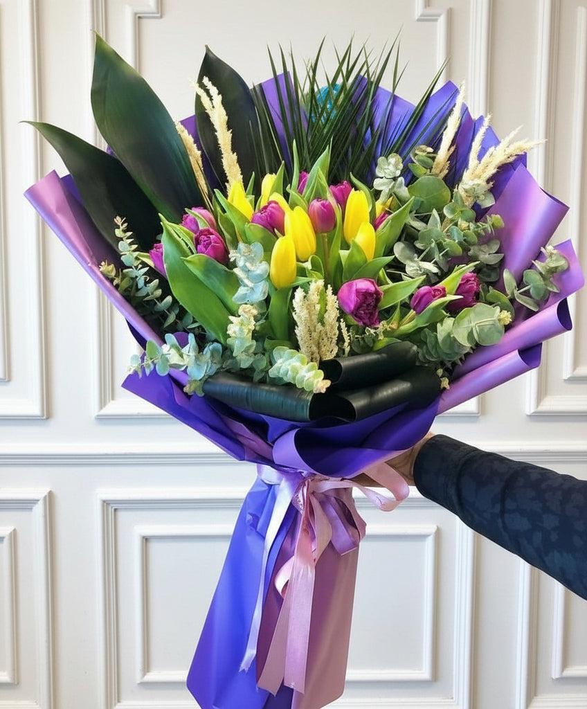 Bouquet of mix flowers & yellow tulips with purple and yellow ribbons held by a person in a floral shop.