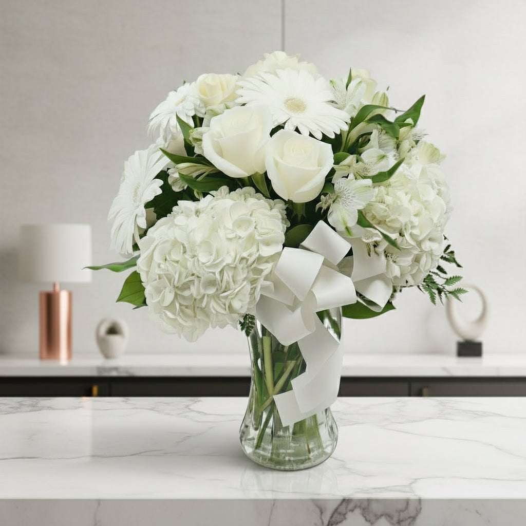 Bouquet of white flowers with green leaves in a clear vase on a marble surface.