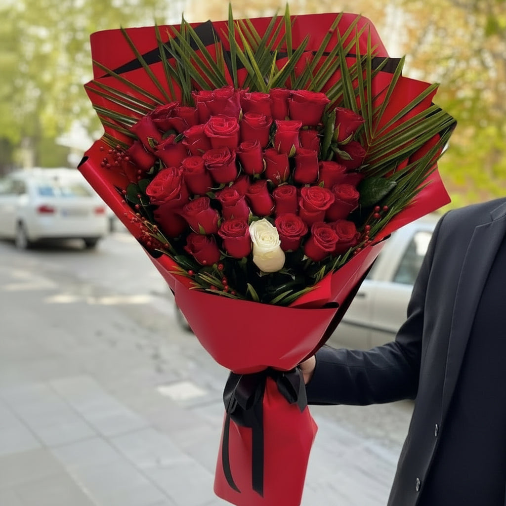 A large bouquet of red roses wrapped in red paper with green leaves, held by a person wearing a dark suit jacket outdoors on a street.