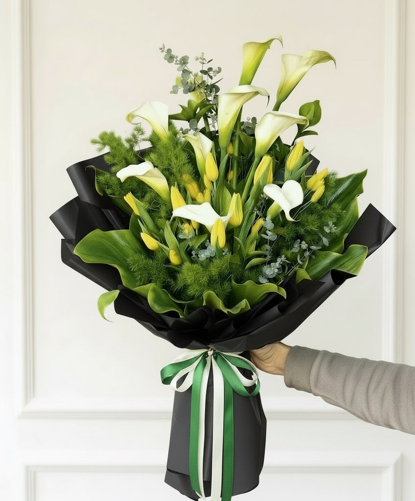 Bouquet of white and yellow flowers wrapped in black paper with a green ribbon, held by a person against a white background.