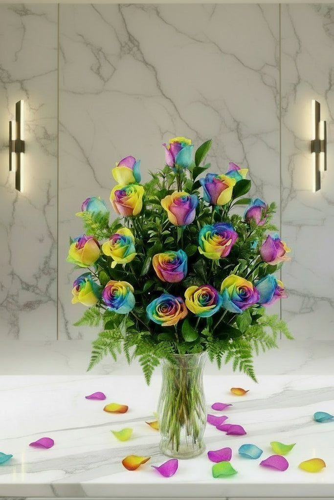 Bouquet of rainbow roses in a clear vase on a marble surface with scattered rose petals.