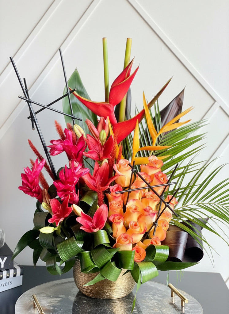 A colorful floral arrangement featuring a variety of tropical flowers such as Roses, Lilies, and Bird of Paradise in a gold-colored container.