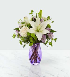 A fresh flower arrangement featuring cream roses, lavender Peruvian Lilies, white Asiatic Lilies, green button poms, lavender statice, bupleurum, and lush greens in a purple vase.
